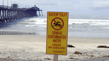Yellow warning sign on beach stating 'Keep Out: Sewage Contaminated Water' with ocean waves and pier in background.