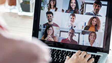 Laptop with a virtual meeting room displaying nine different people, in the front someone's back and hand