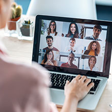 Laptop with a virtual meeting room displaying nine different people, in the front someone's back and hand