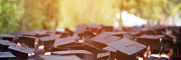 Group of graduates wearing black caps with red tassels at an outdoor graduation ceremony, seen from behind
