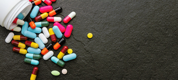 Image features a collection of multicoloured pills spilling out from container, on black counter.
