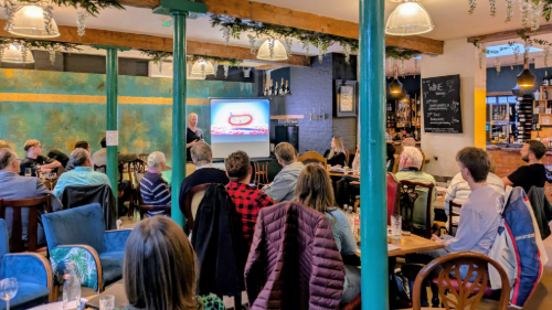 A group of people seated in a bar attentively watching the presenter near a screen displaying a bacteria diagram
