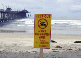 Yellow warning sign on beach stating 'Keep Out: Sewage Contaminated Water' with ocean waves and pier in background.