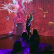 Host interacting with bacteria projection in a colourful, immersive digital room while an audience watches